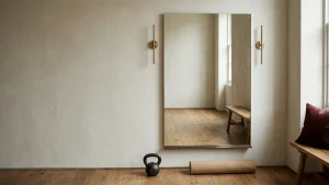 Large frameless gym mirror wall on bone plaster, flanked by two slim brass sconces at face height, with a kettlebell and rolled mat below
