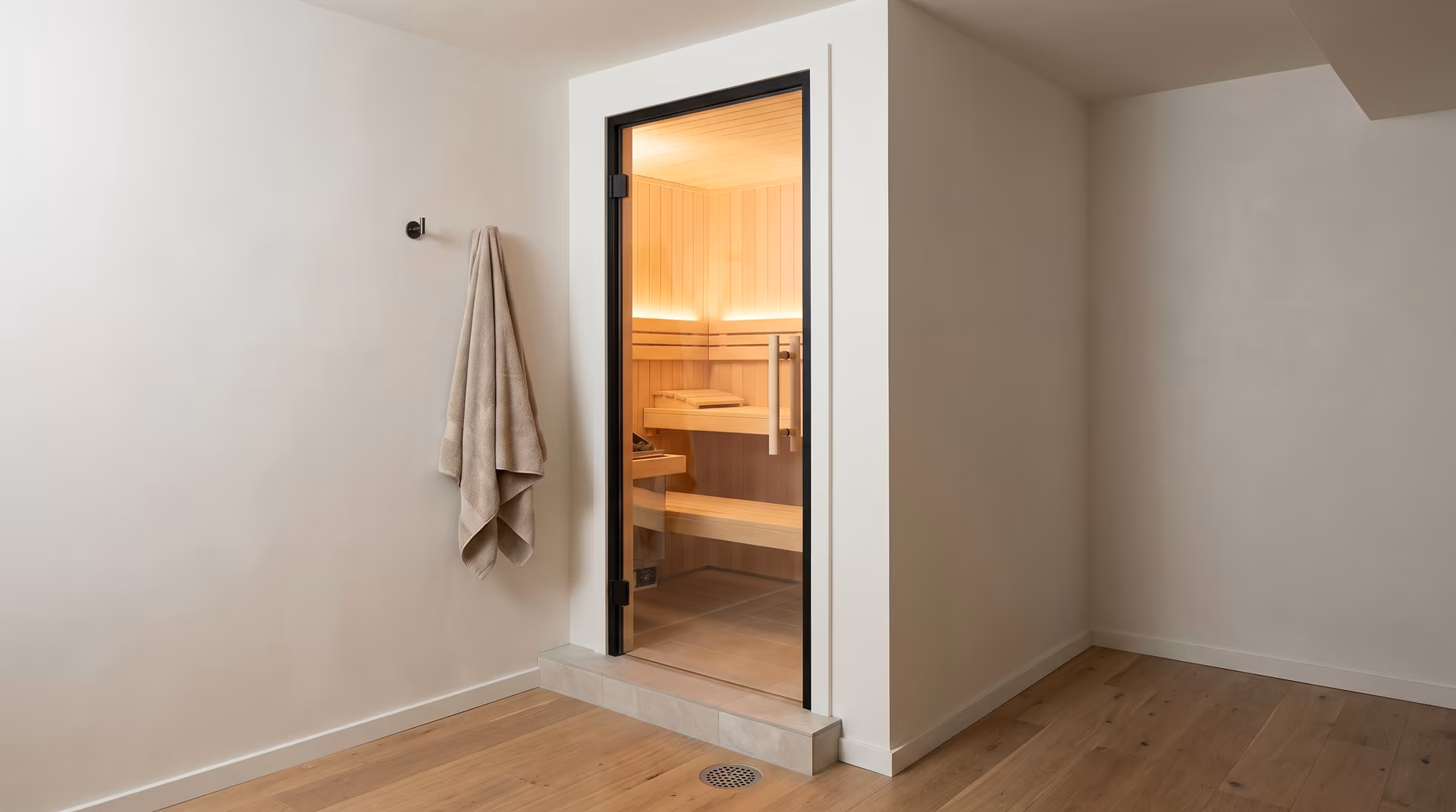 Compact sauna cabin with a glass door built into the corner of a plain white room with oak flooring and a towel on a hook