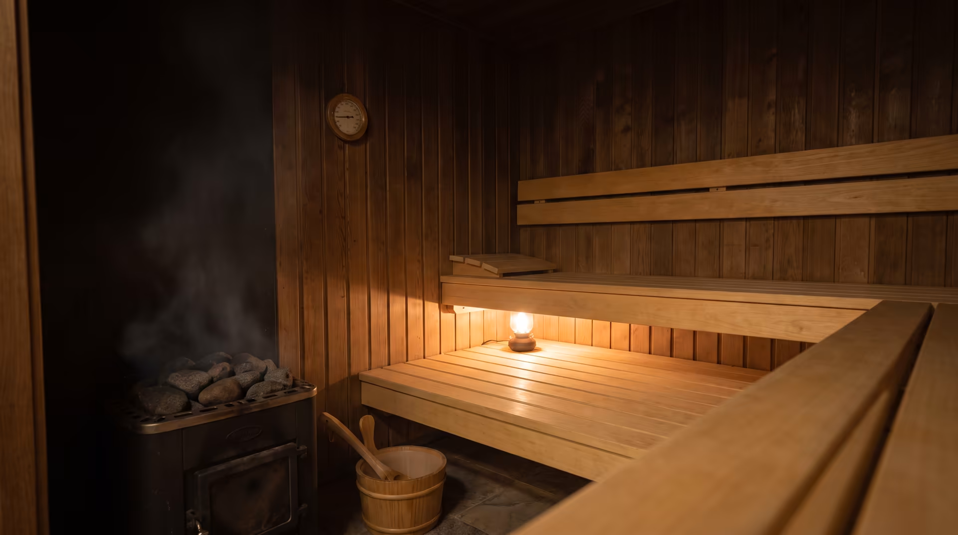 Interior of a traditional wood sauna with tiered cedar benches, a wood-burning stove filled with stones, and a bucket and ladle