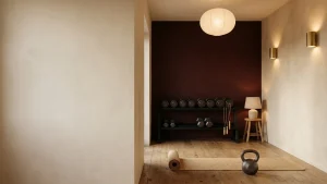 Home gym lit in the evening by a diffused ceiling fitting, two brass wall sconces and a low table lamp, with a deep oxblood accent wall